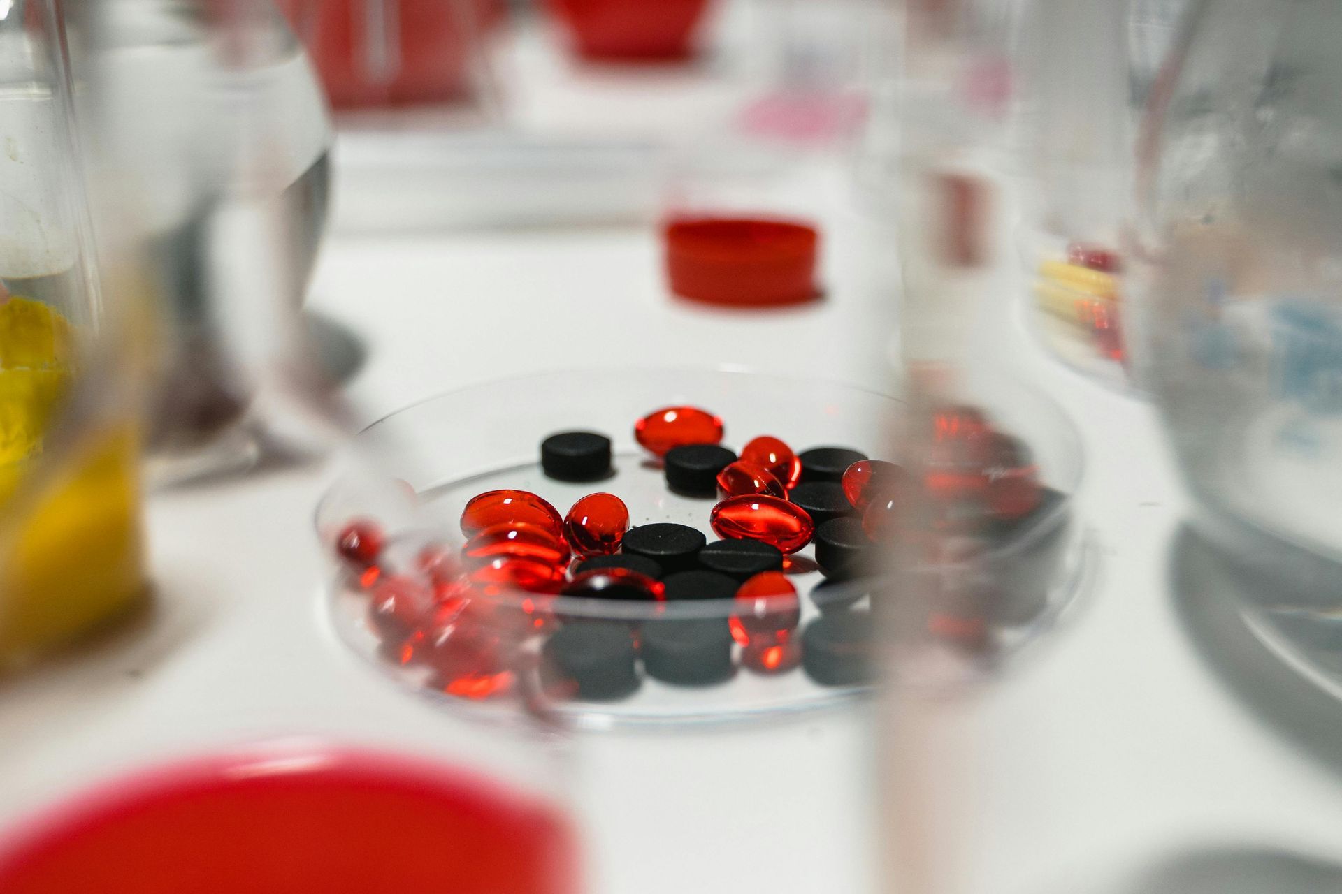 Red and black pills in a petri dish on a white table, surrounded by other laboratory equipment.