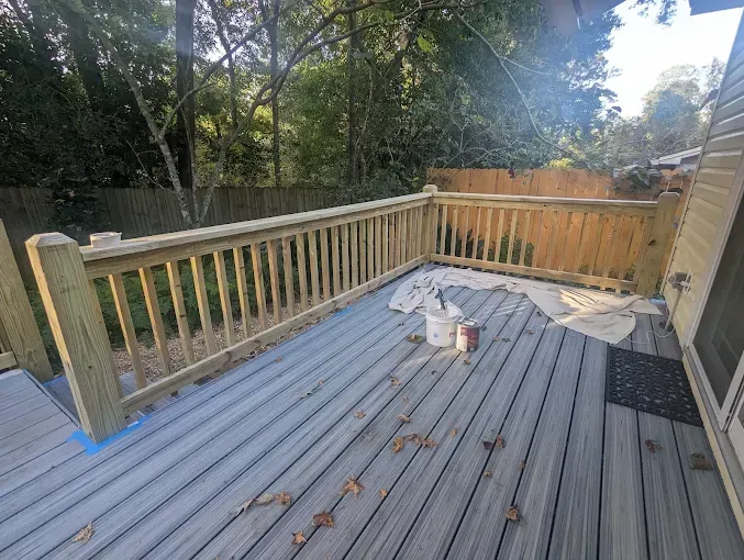 Wooden deck with railing, painting supplies, and trees in background.