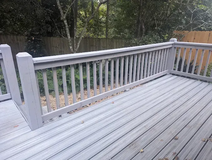 Gray wooden deck with matching railing, looking out onto a yard with trees and a fence.