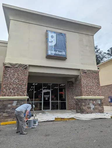 Person painting the entrance of a Goodwill store.  Sign is covered in plastic. Exterior brickwork.