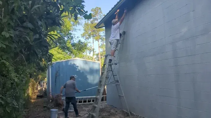 Two people painting the exterior of a light blue building. One on a ladder, other on the ground with a roller.