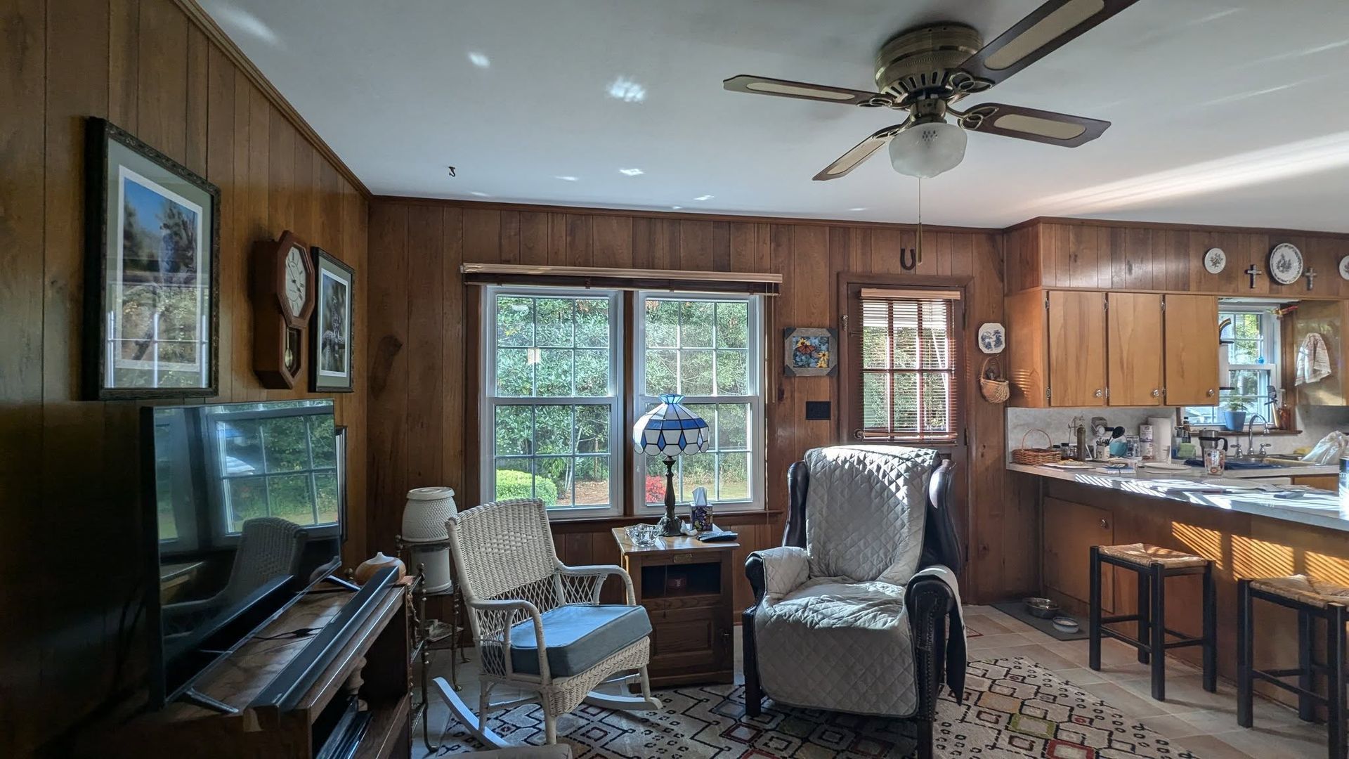 Cozy living room with wood paneling, a rocking chair, and a bar area, with windows looking out at greenery.