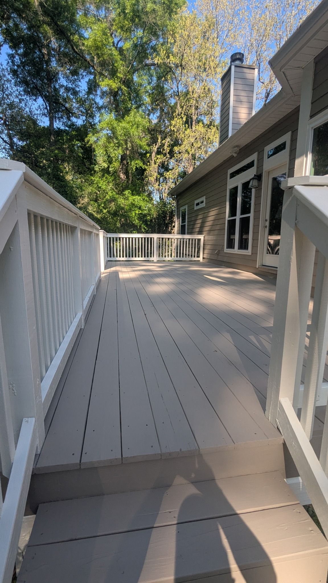 A wooden deck with white railings extends from a gray-sided house, trees in the background.