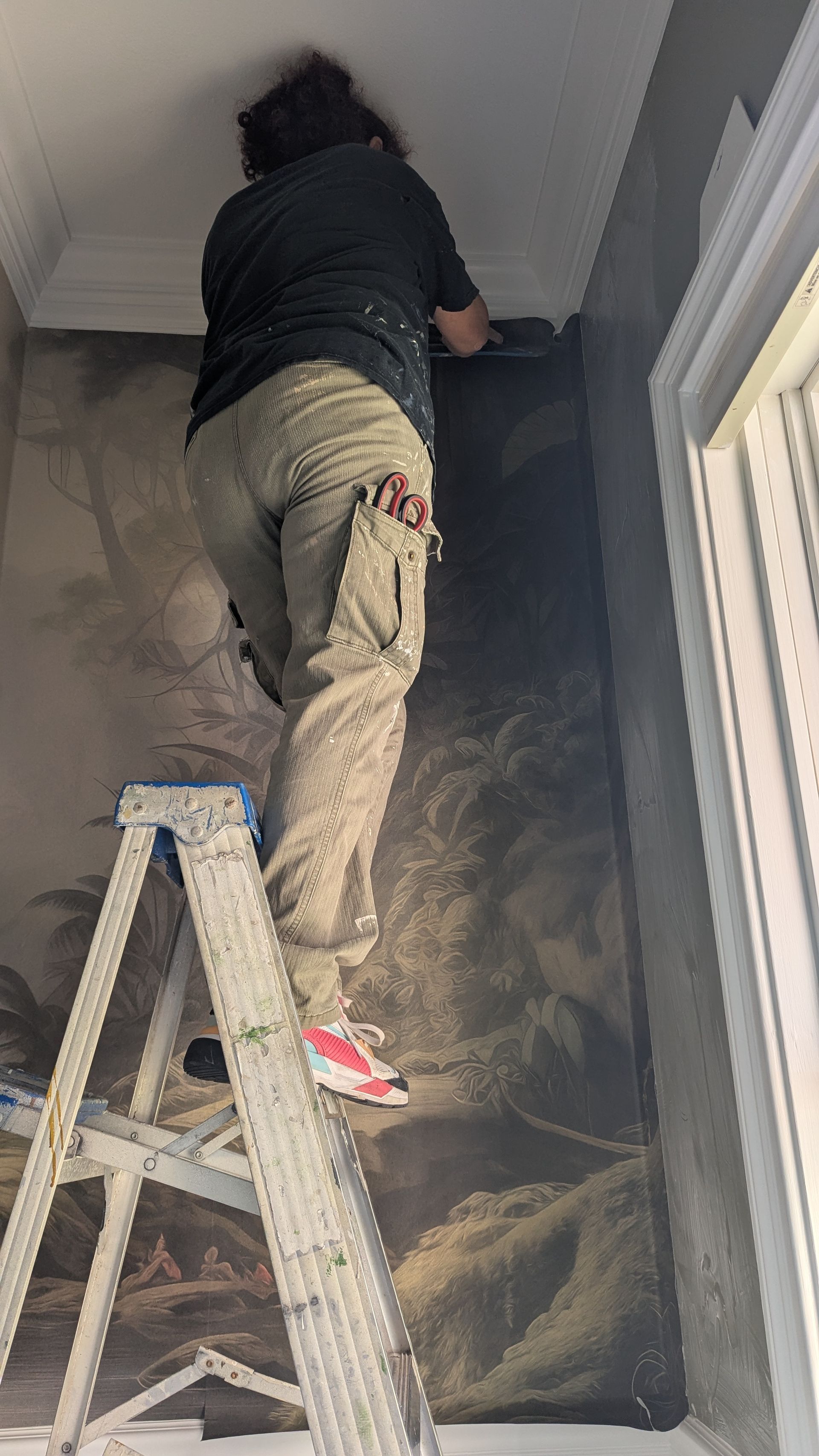 Person on a ladder installing wallpaper in a room. They are wearing work clothes and reaching towards the ceiling.