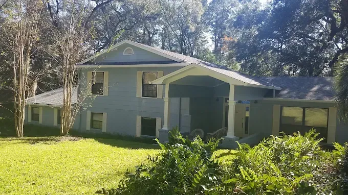 Two-story blue house with white trim, porch, and green lawn surrounded by trees.
