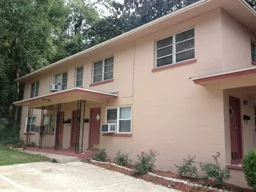 Two-story beige apartment building with red doors and trim, set in a wooded area.