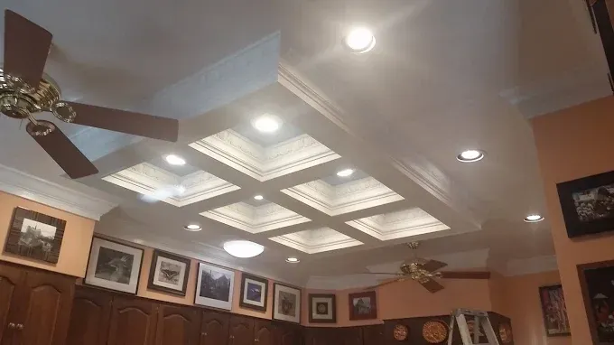 Coffered ceiling with recessed lights and crown molding. Two ceiling fans and artwork on walls.