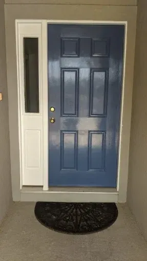 Blue front door with sidelight, white trim, and gray frame. Black doormat on concrete.