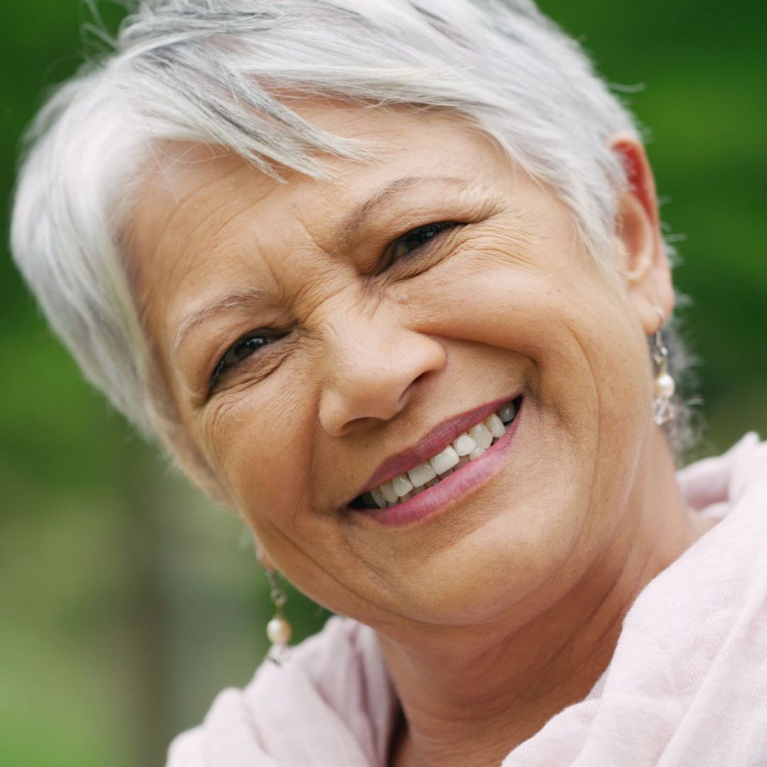 Smiling older woman with short, white hair and light pink clothing, outdoors.
