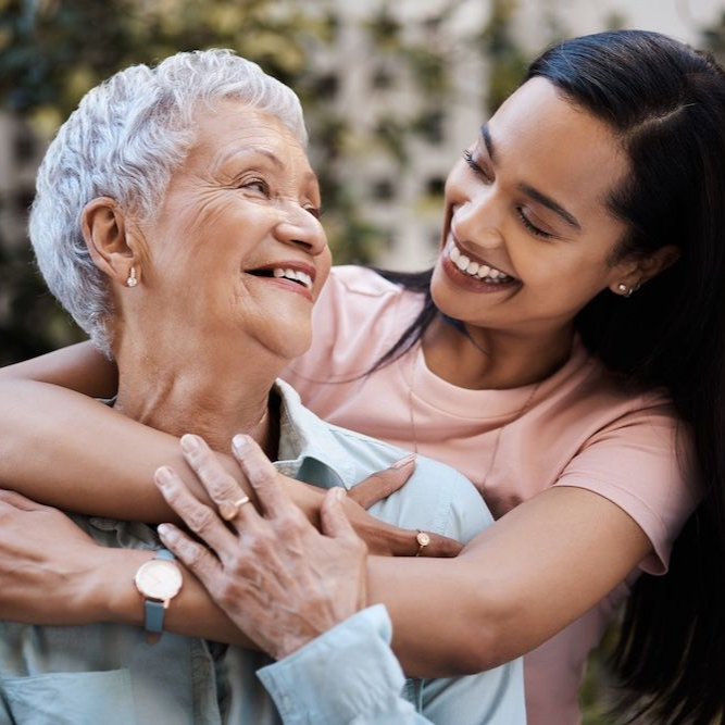 Woman embracing and smiling at an elderly woman, both outdoors in a garden, soft colors.