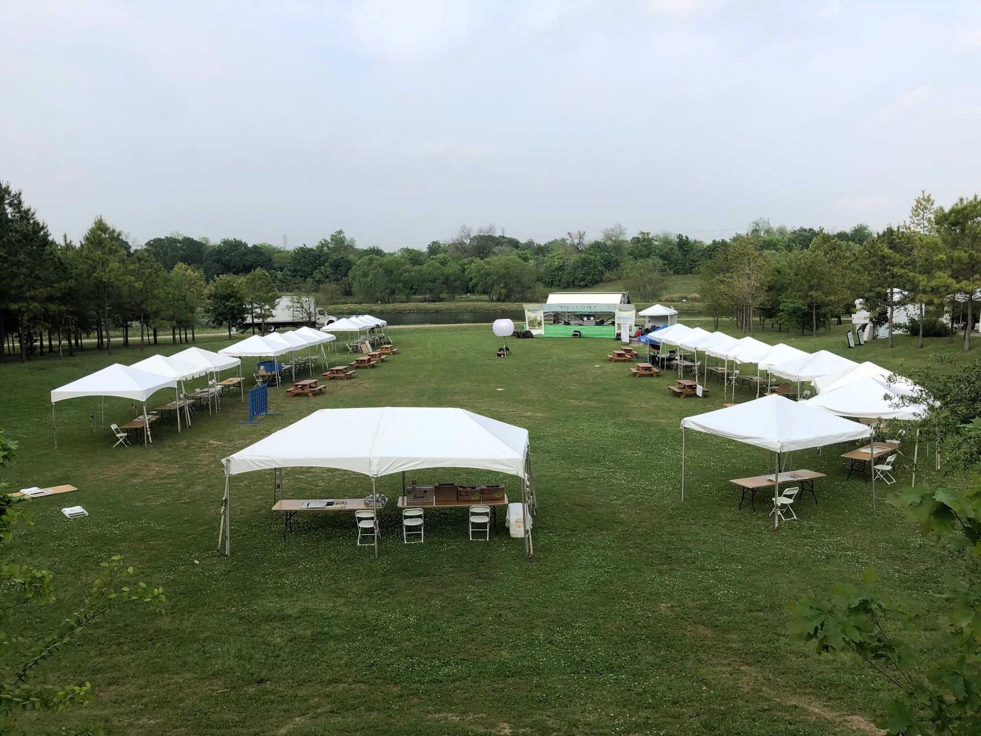 Outdoor event with white tents, tables, and chairs on green grass. Trees and pond in the background.