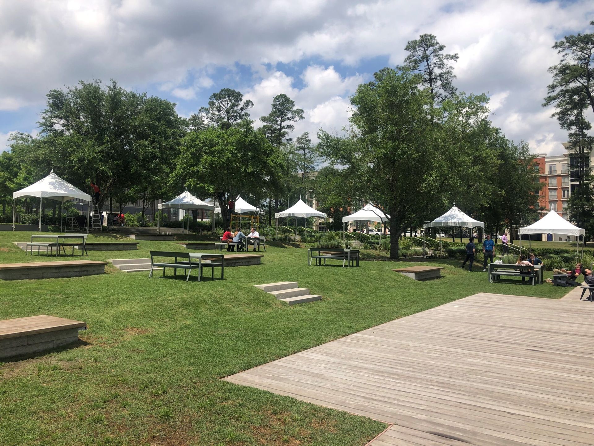 Park with picnic tables, white tents, and people under trees. Cloudy sky overhead.