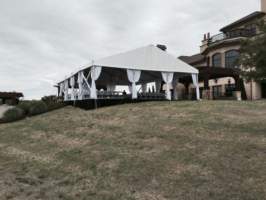 A large white tent with sheer curtains is set up on a grassy hill next to a large house.