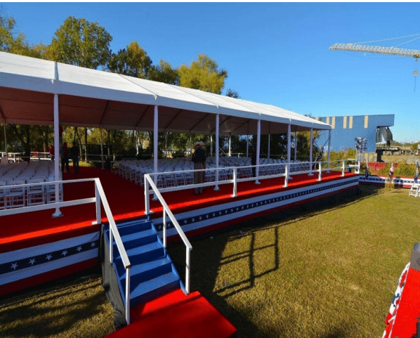 A red-carpeted stage with white chairs under a canopy, decorated with red, white, and blue, outdoors.