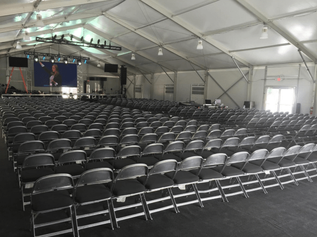 Rows of black chairs face a stage with a screen inside a large tent.