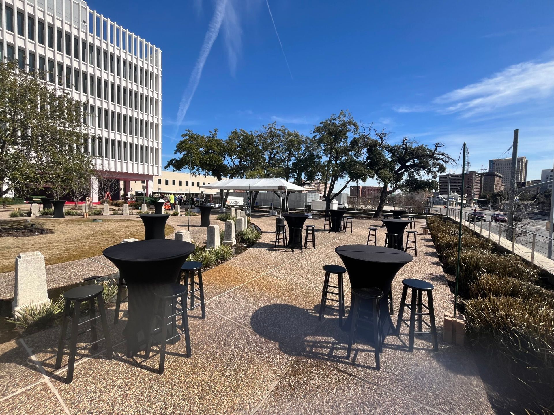Outdoor event space with black high-top tables and stools. White building, trees, and blue sky in background.