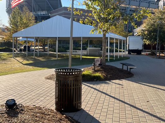 Large white tent set up in a park on a sunny day. A trash can and trees are in the foreground.
