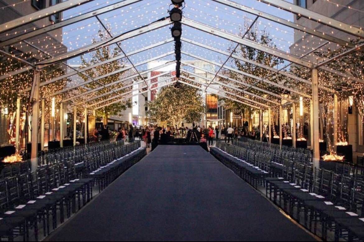 Fashion runway under a clear tent; rows of chairs, black carpet, trees, and people.