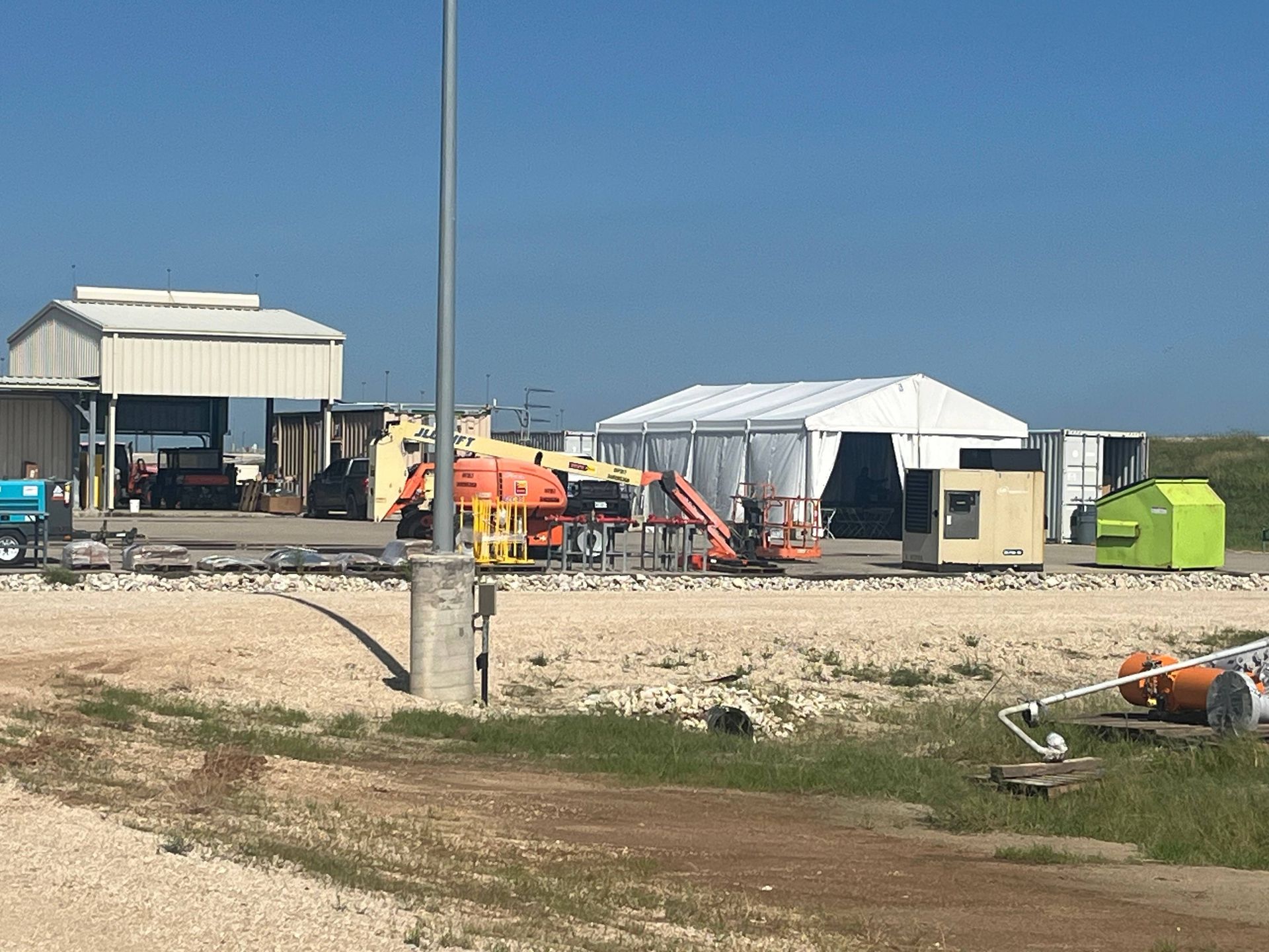 Construction site with vehicles, white tent, and buildings under a blue sky.