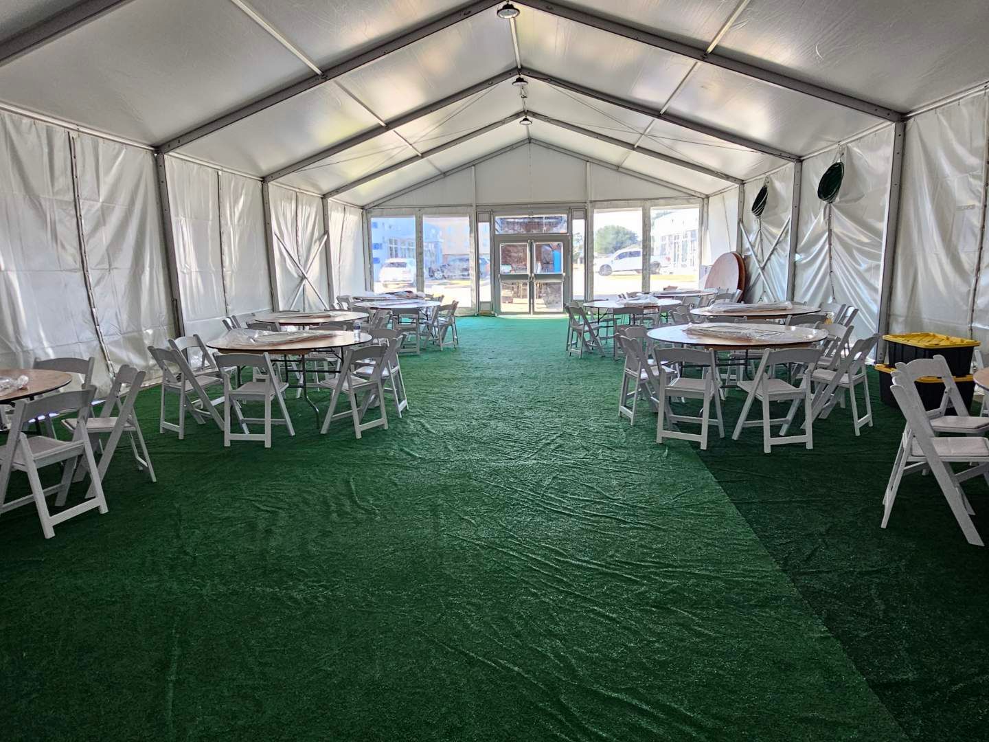 A large tent interior with round tables and chairs on green turf, sunlight streaming through doors.