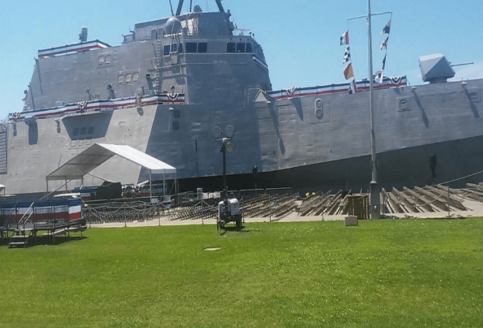 Large gray naval ship docked on green grass, sunny day. Flags and construction scaffolding visible.