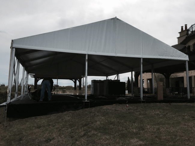 White tent set up on grass, with a person visible underneath, and a building in the background.