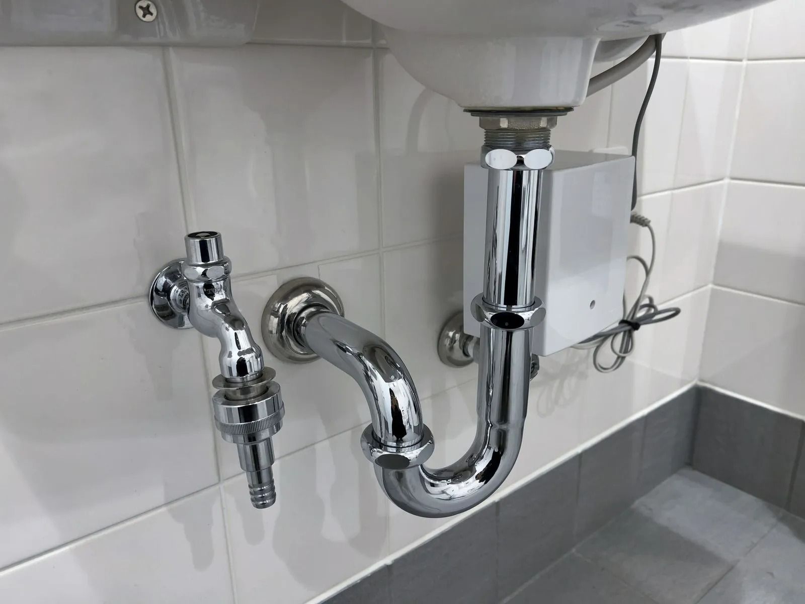 Under-sink chrome plumbing with P-trap and shutoff valve against white tiled wall