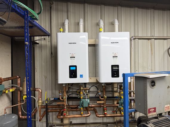 Two white wall-mounted boilers with copper piping in an industrial utility room.