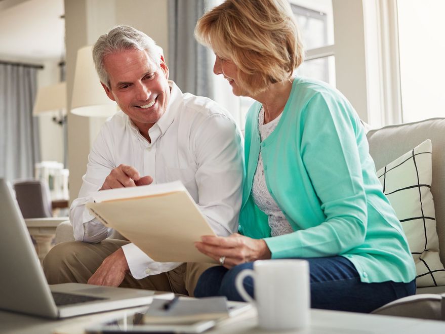 Couple on couch reviewing papers, man points, laptop and mug on table, sunny living room.