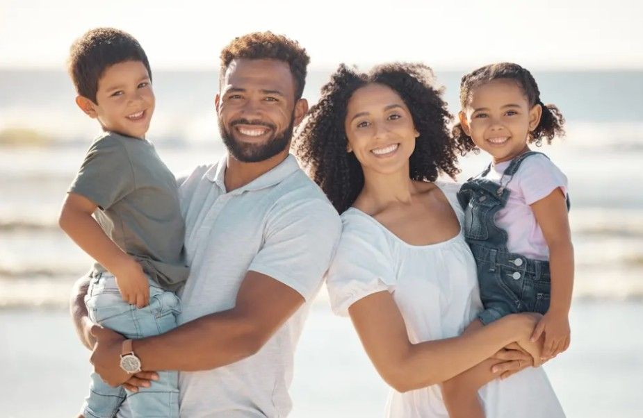 Family of four smiling on a beach. Man holds boy, woman holds girl, all facing camera.