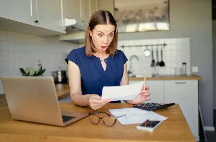 Woman with surprised expression looking at bills in a kitchen, laptop and calculator nearby.