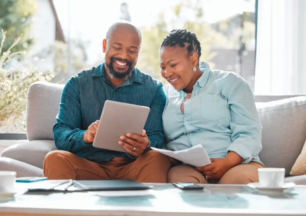 Couple on a couch looking at tablet and papers; smiling, light-filled living room.