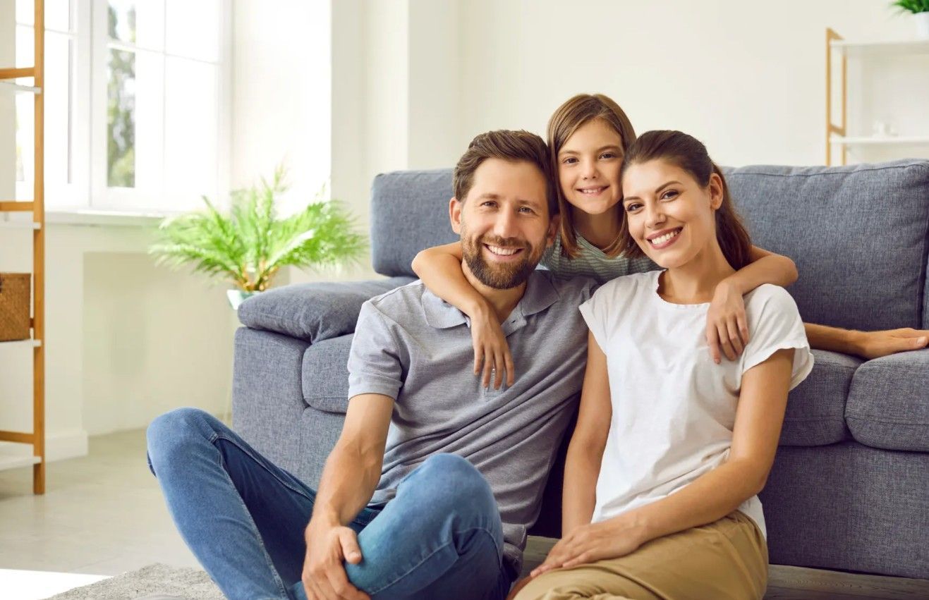 Family of three smiling, posing on the floor in front of a blue couch. Daughter hugs parents. Bright living room.