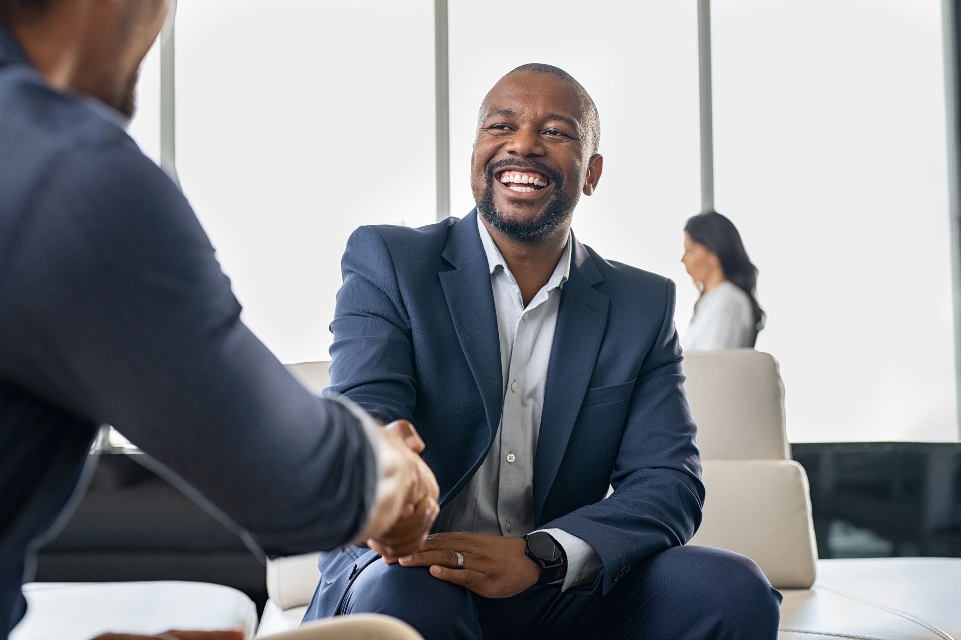 Man in a suit smiles, shaking hands with another person in a well-lit office setting.