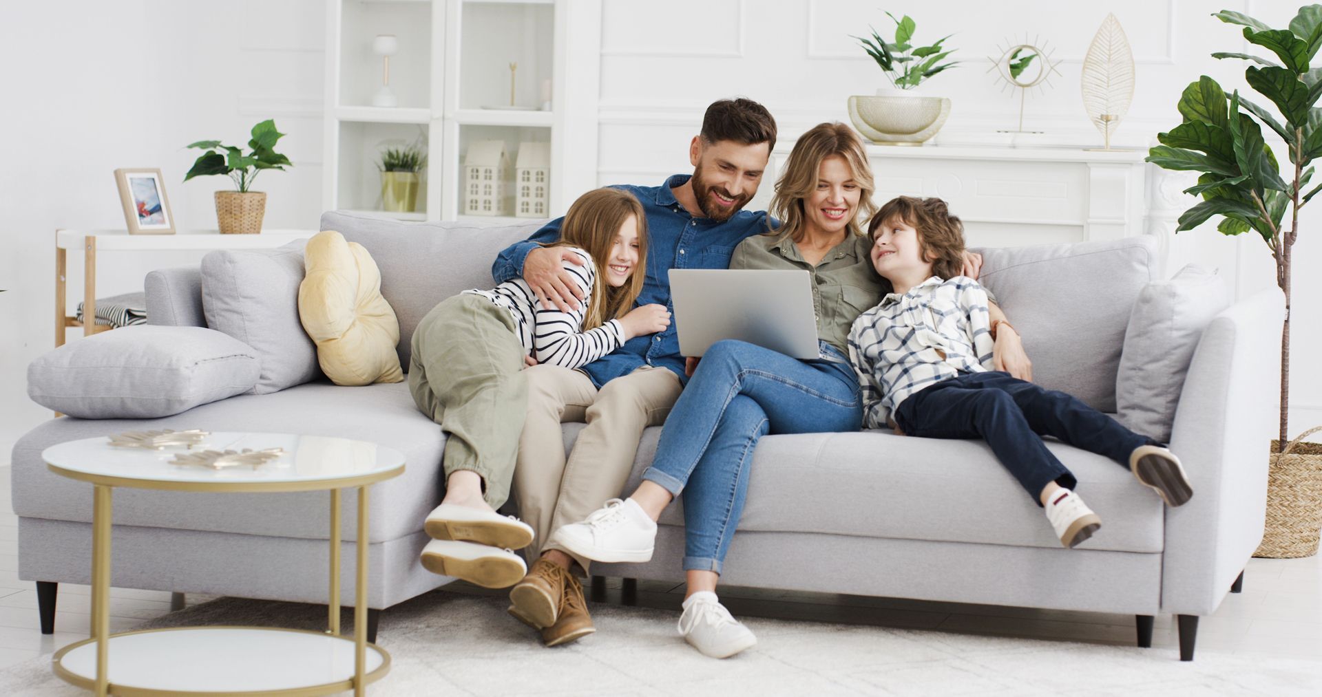 Family of four smiles while looking at a laptop on a gray couch in a living room.