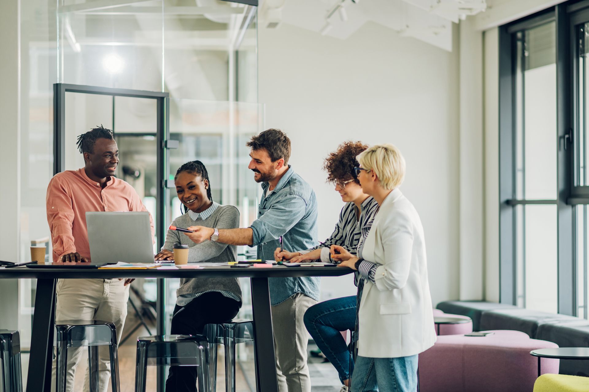 People collaborate at a high table, looking at a laptop and documents in a bright office.