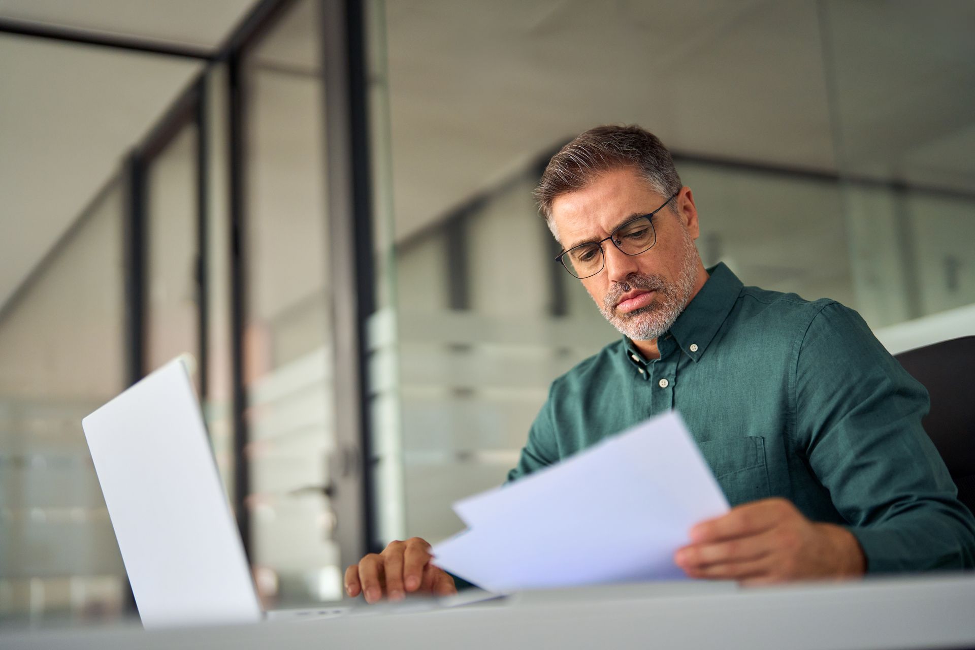 Man in glasses reviewing paperwork at a desk with a laptop, in an office.