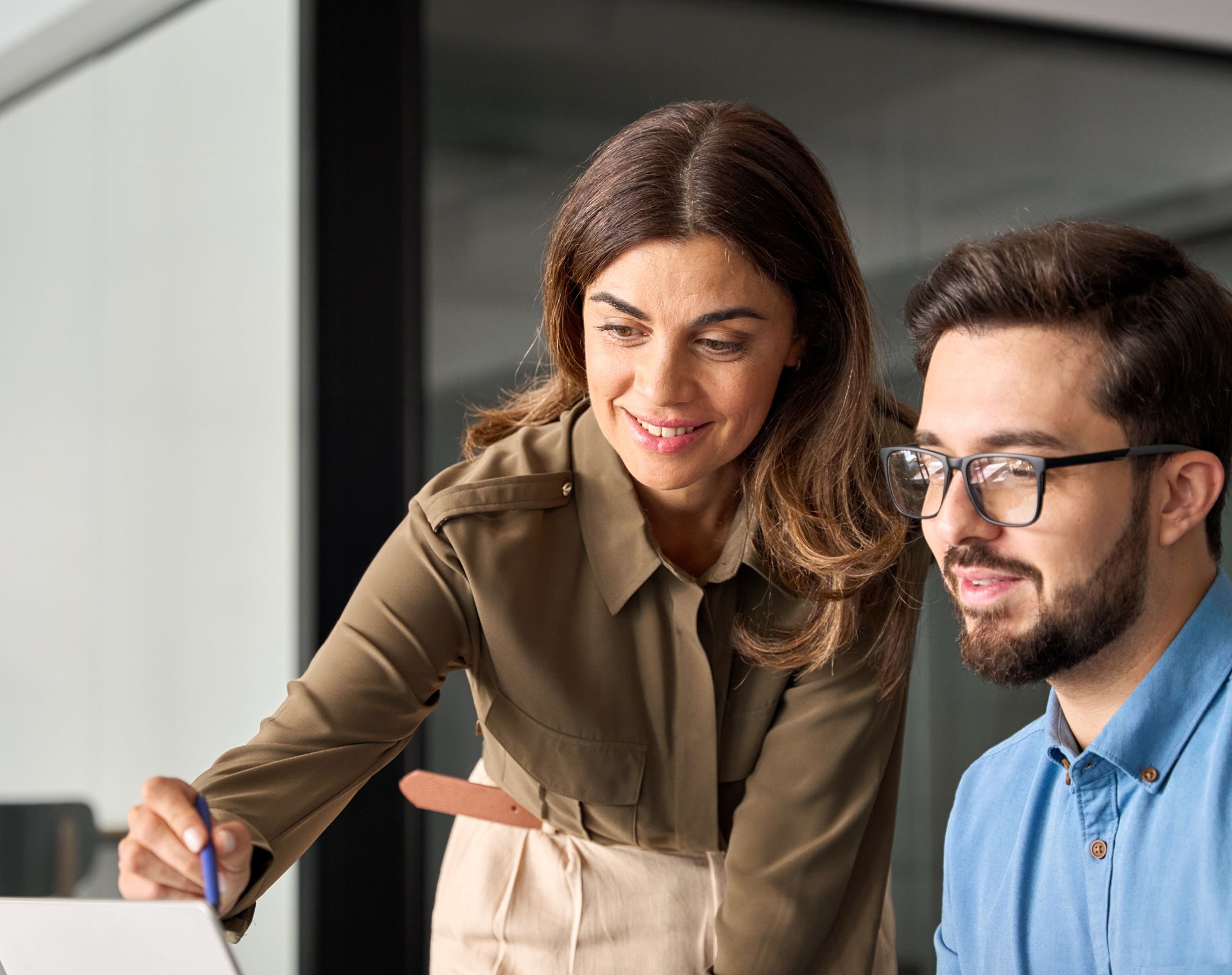 Woman pointing at laptop screen, smiling at man wearing glasses, office setting.