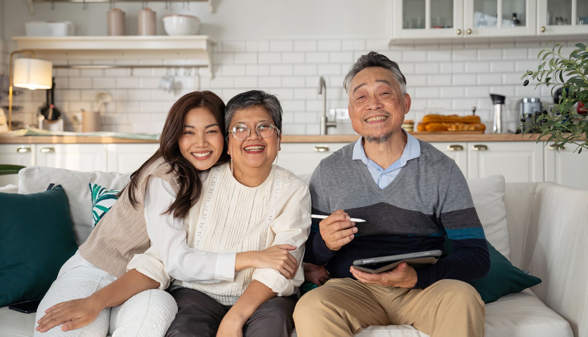 Family of three smiling on a couch, holding tablet, indoors, in front of a white tiled wall.
