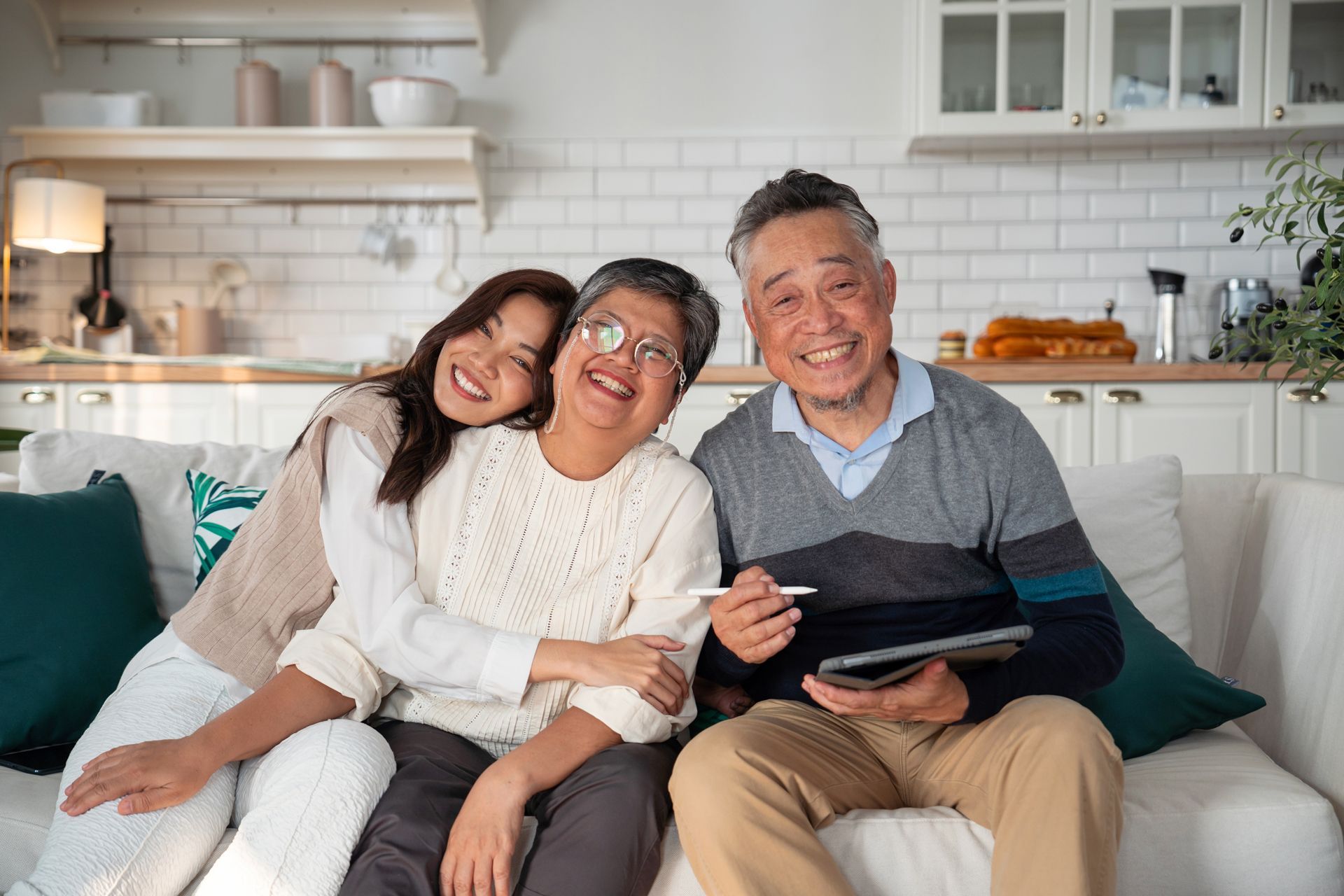 Woman hugs smiling elders on a couch in a bright living room, holding a tablet.