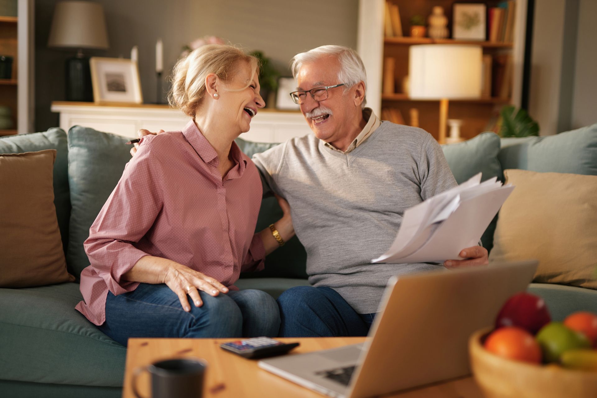Smiling couple on a sofa reviewing documents, laptop open on coffee table.