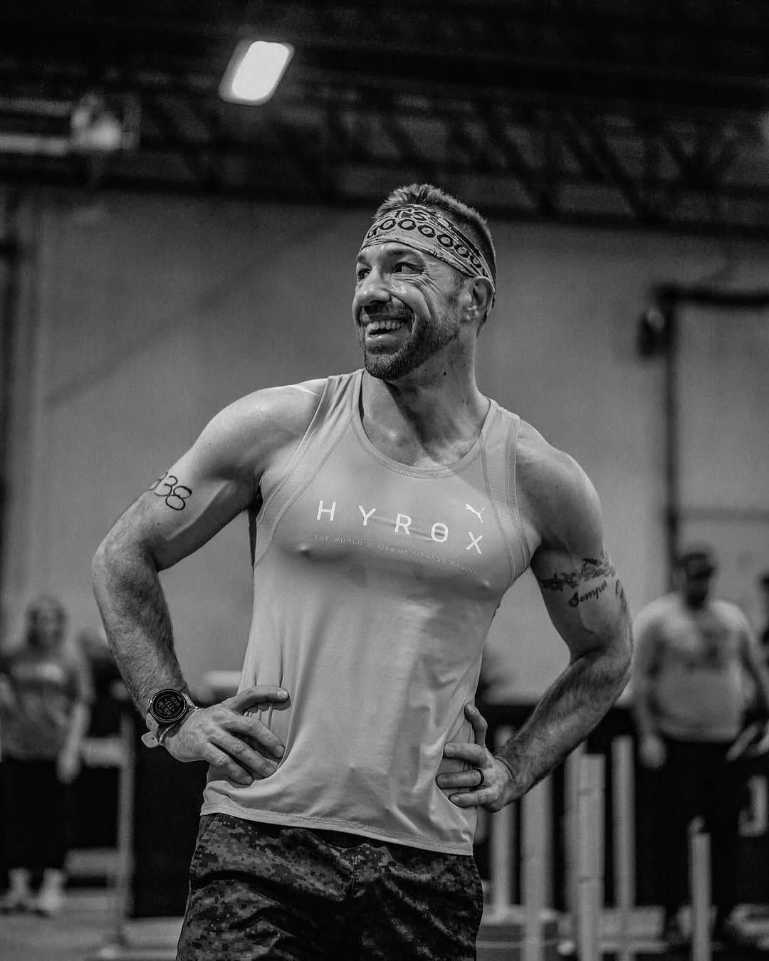 Un homme portant un débardeur et un bandeau se tient debout dans une salle de sport, les mains sur les hanches.