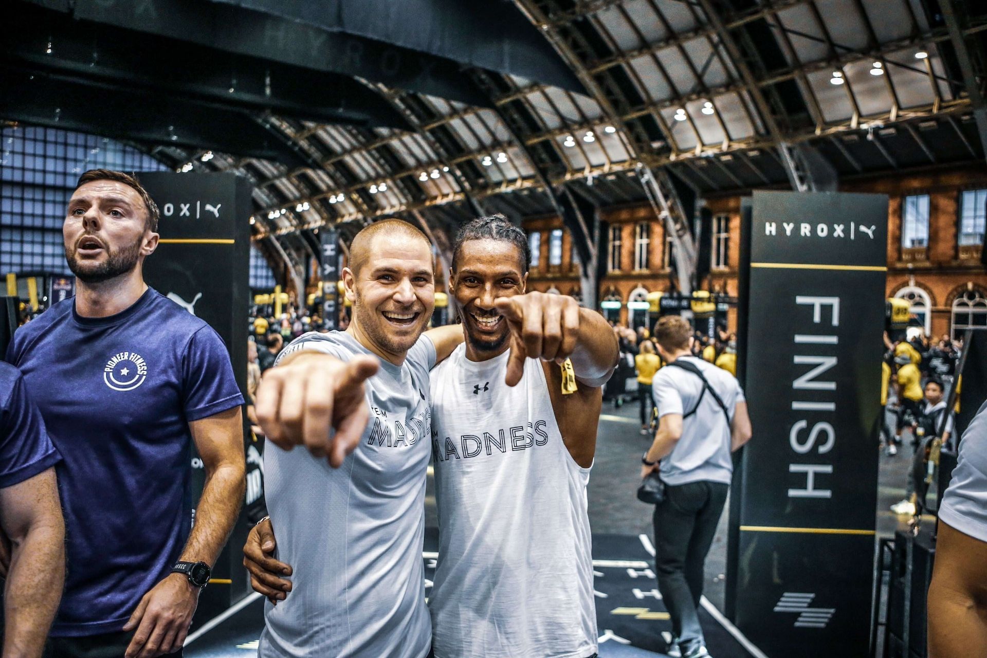 Deux hommes se tiennent l'un à côté de l'autre dans une salle de sport et pointent du doigt la caméra.