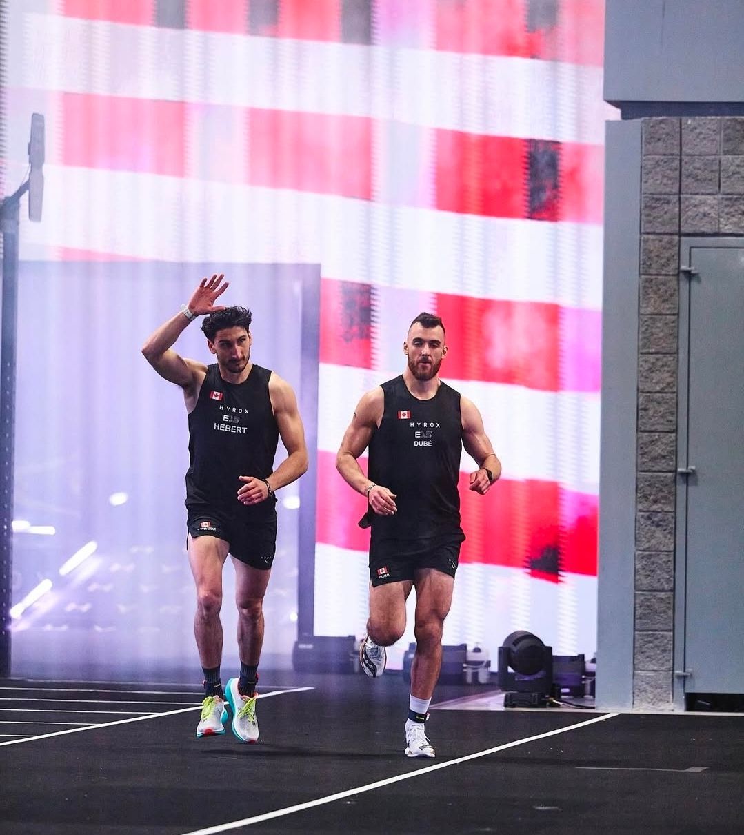 Two athletes in black tank tops and shorts run towards the camera on an indoor track with a bright red-and-white background.