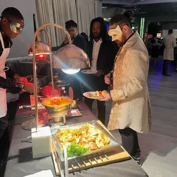 A man wearing a mask is standing at a buffet table holding a plate of food.