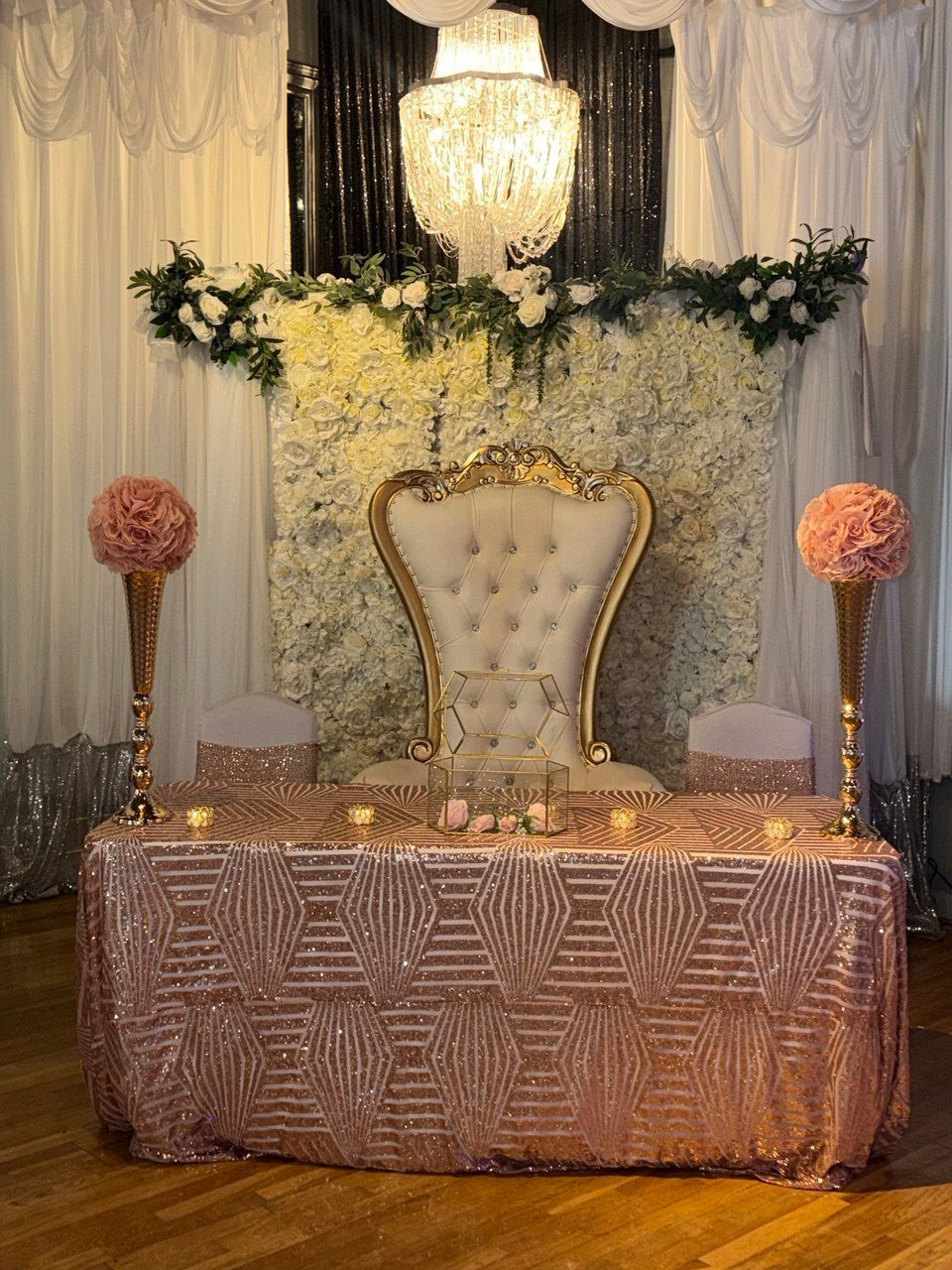 Elegant event backdrop with gold tufted chair, floral arch, chandelier, and pink-draped table