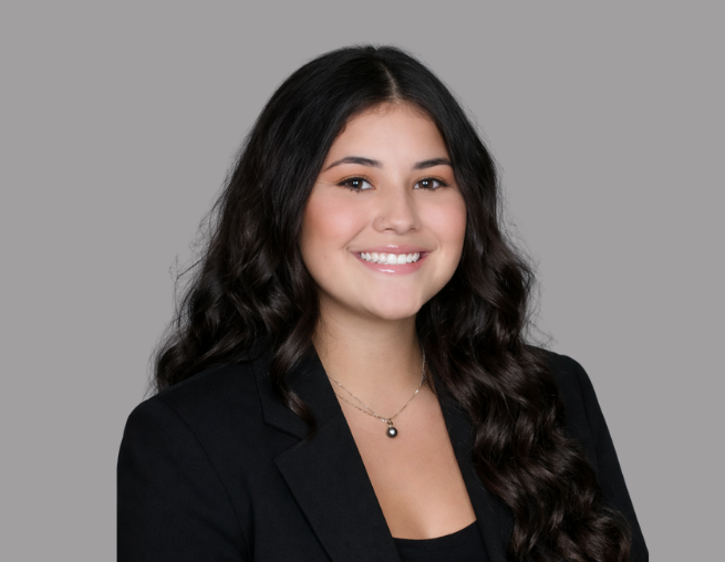Woman with dark hair in a black blazer smiling against a gray background.