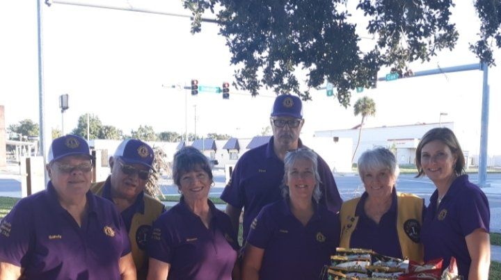 Group of seven people in purple shirts and vests, some wearing hats, outdoors.