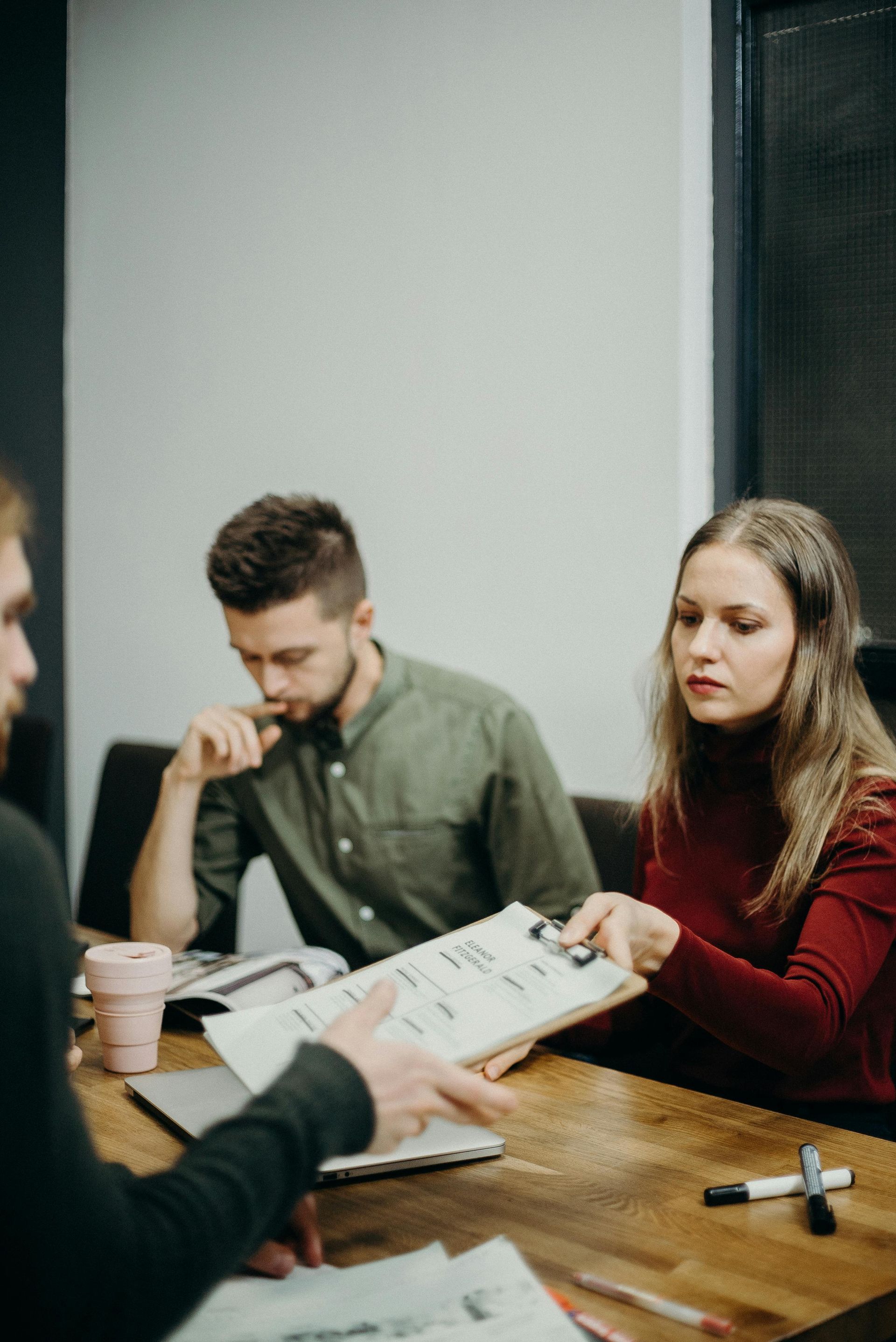 People at a table review documents. Woman in red sweater holds a clipboard; man in green shirt looks on.