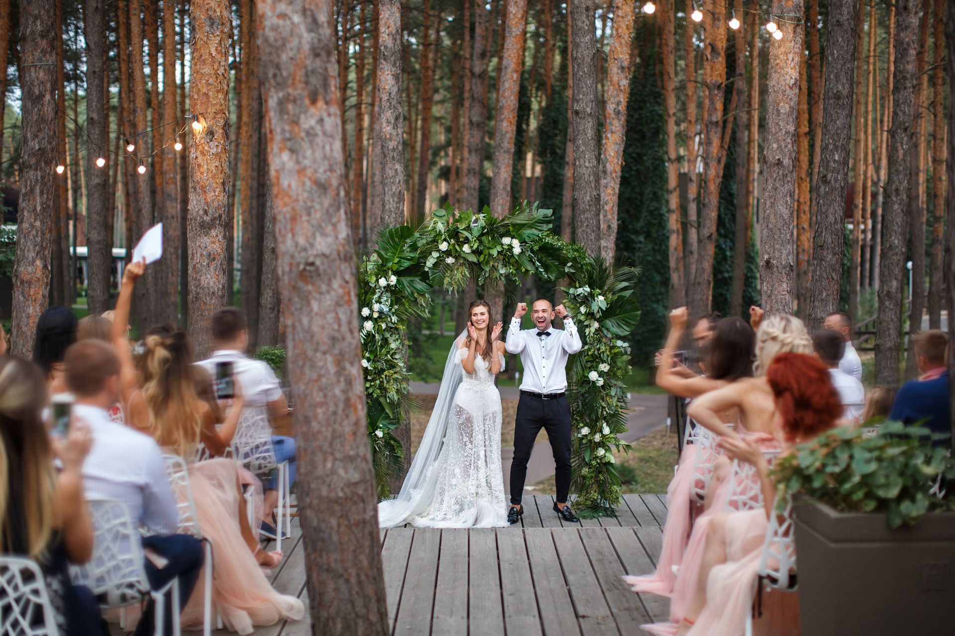 Newlyweds celebrate under a floral arch in a forest. Guests cheer, some hold phones, wooden platform.